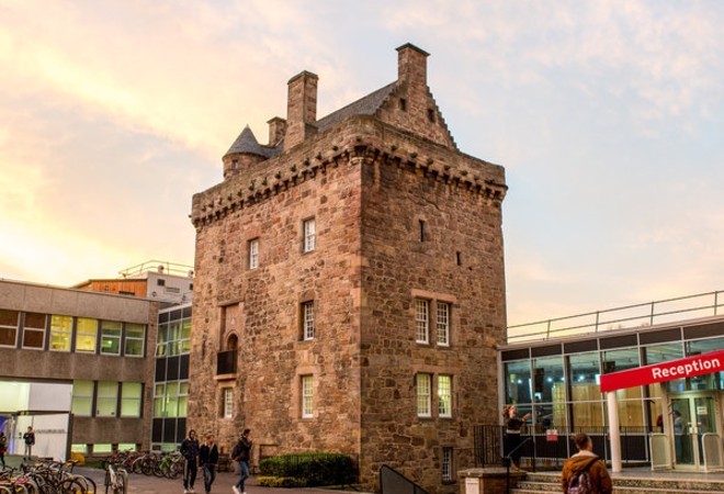 "Old university building with reception sign to right hand side set against lightly clouded sky"