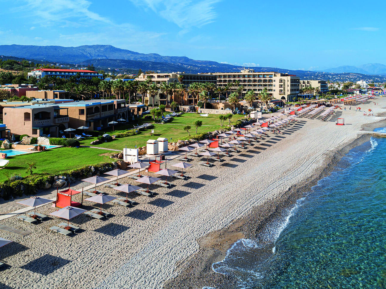 Aerial view above the sea of the beach with many sun loungers and umbrellas along the sand with the hotel complex directly behind with a lawn and the buildings behind.