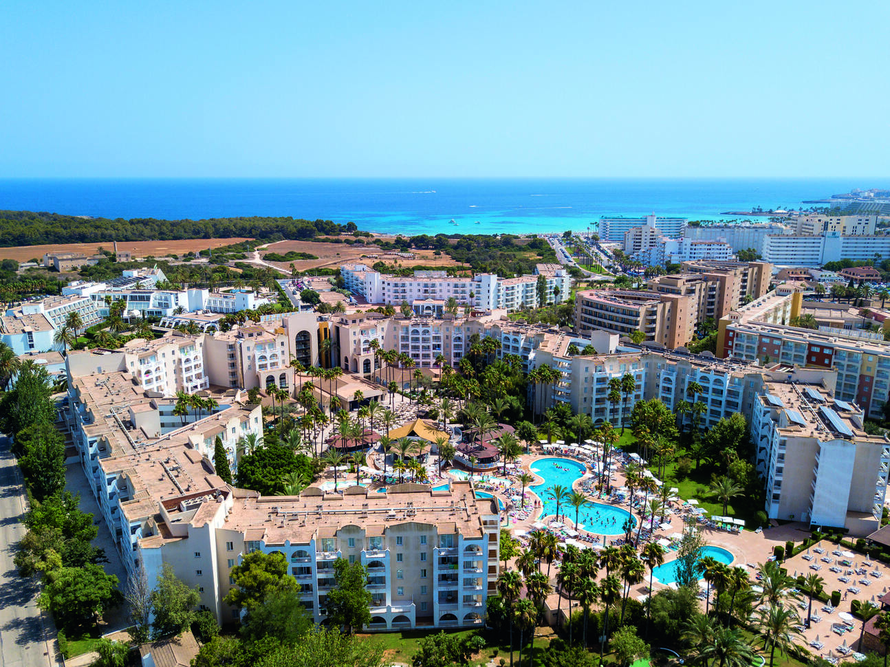 Aerial view of the large hotel complex with 3 different pools, many sun loungers, people dotted around on the loungers and in the pool and many palm trees. The hotel complex is surrounded by many more hotel buildings with the blue sea in the background on a clear sunny day.