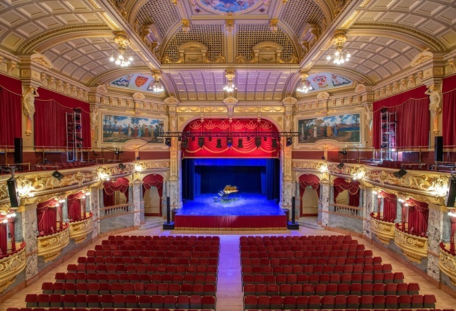 "A photograph of one of Harrogate Convention Centre's exhibition spaces.  This is a beautifully decorated and ornate room of reds and golds with artwork in the ceiling.  A centre stage with red drapes is set before rows of seating with VIP boxes set to each side"