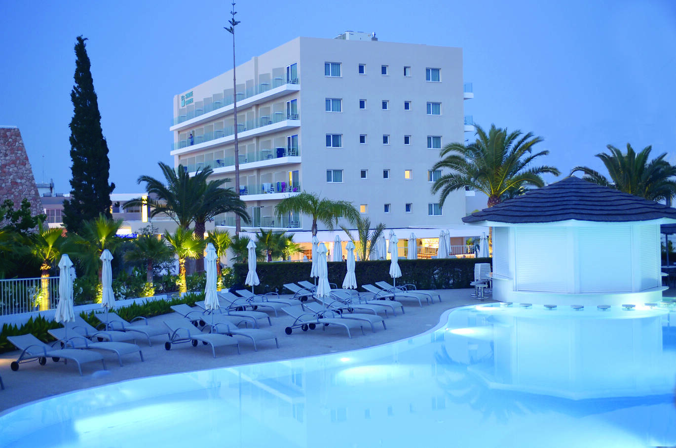 Pool side view in the dusk of sun loungers on the pool side and the swim up bar all closed up. With palm trees behind and part of the tall hotel building behind.