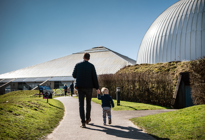 "Photo of outside the centre, there is a grey dome shape building to the right and a grey triangular roof building straight ahead. There is a man and a boy walking up the path towards the building."