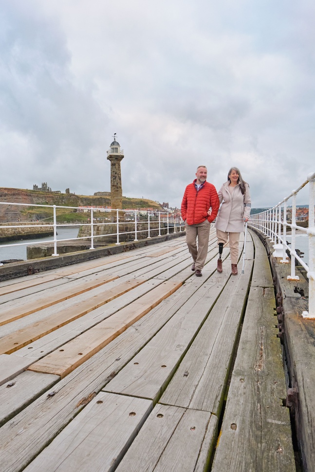 "Two people walking along a pier with a lighthouse in the distance"