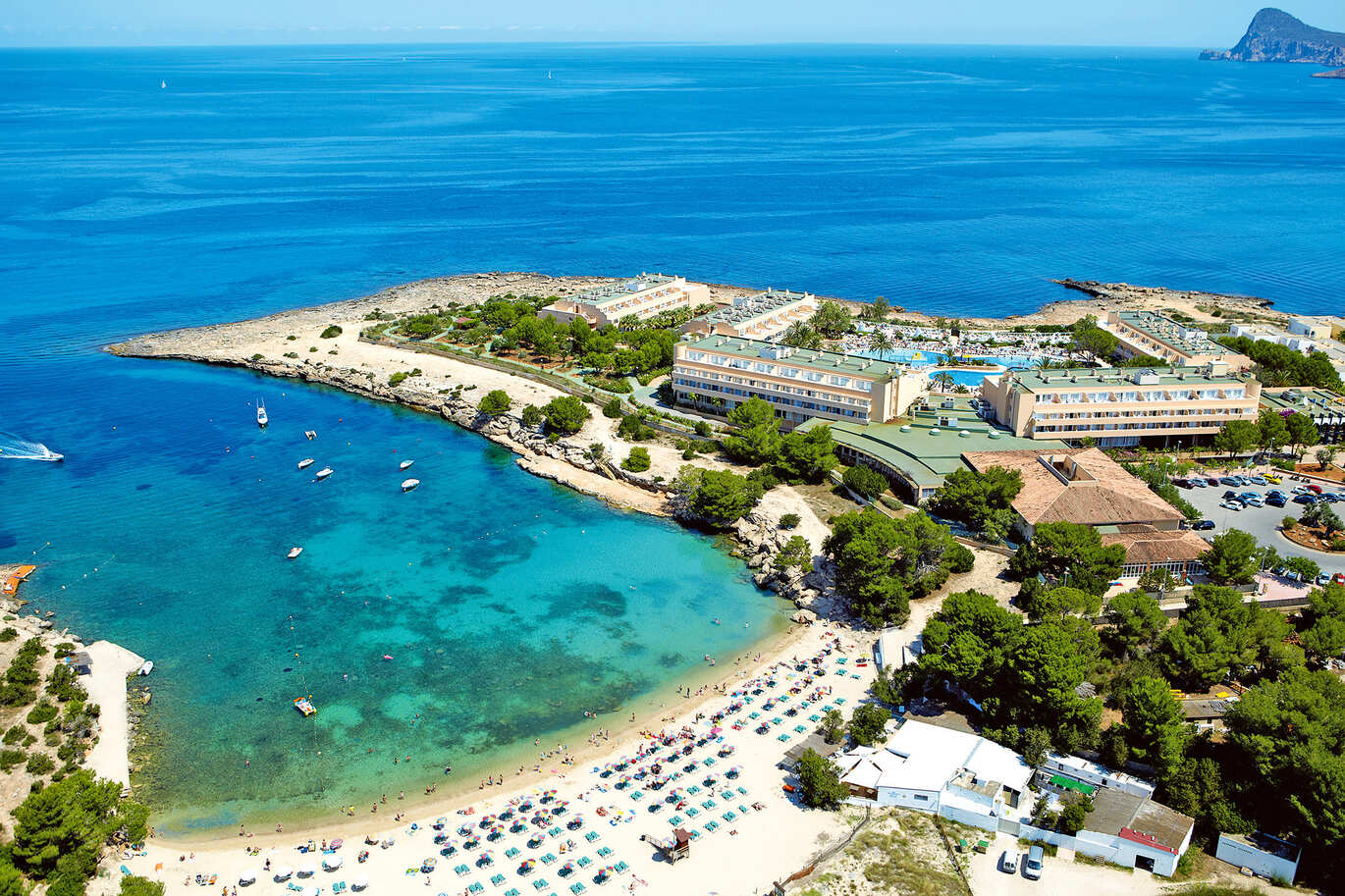Beautiful aerial view of the hotel and sea with a few boats in and a lovely white beach with lots of people and sun beds. You can see the hotel complex with a big pool and the car park just next to the hotel buildings with lots of green trees surrounding it all. 