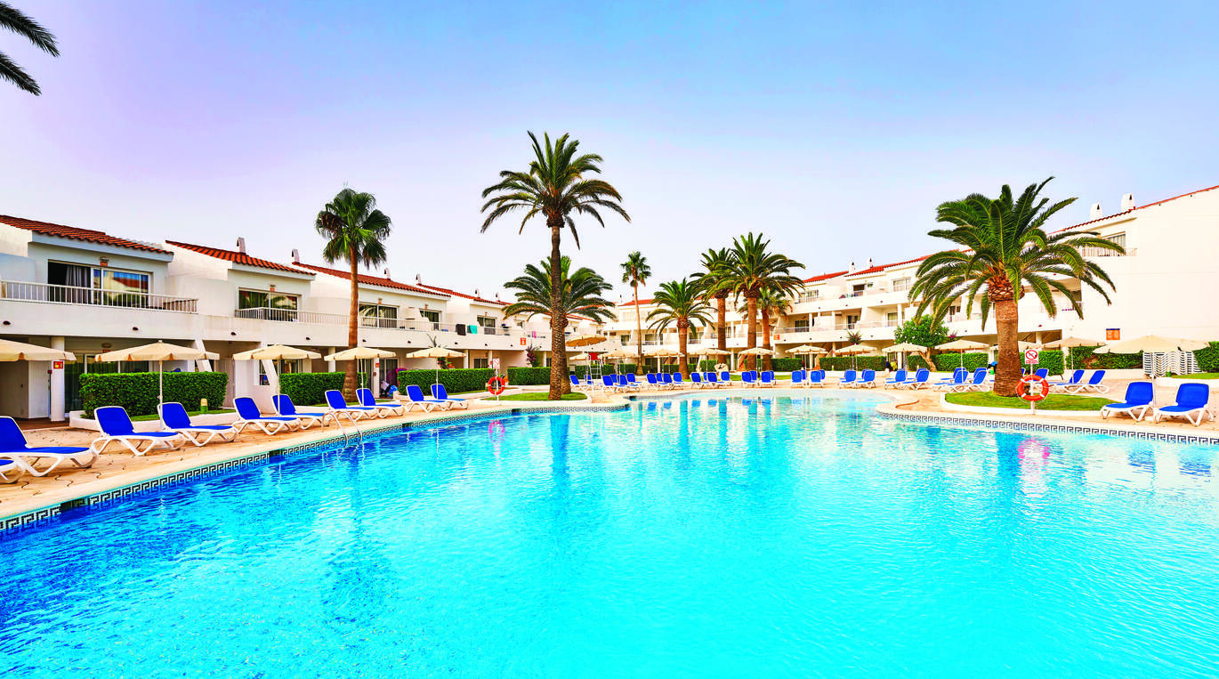 A view of the large, blue swimming pool surrounded by many blue sun loungers and white umbrellas with palm trees around. Behind this is the hotel building where you can see a cluster of hotel rooms with balconies. The sky above is clear and blue with a slight purple tinge.