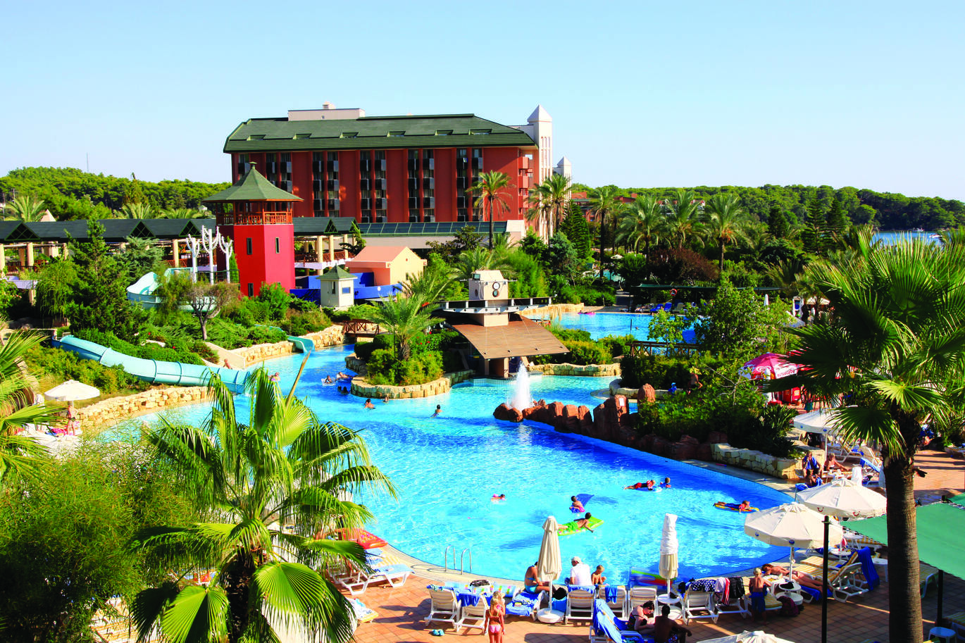 View of the pool area surrounded by greenery and trees with the hotel building in the background and the blue sky above.