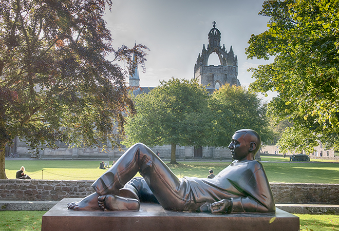 "Sculpture of man lying down in foreground with building showing above trees in background"