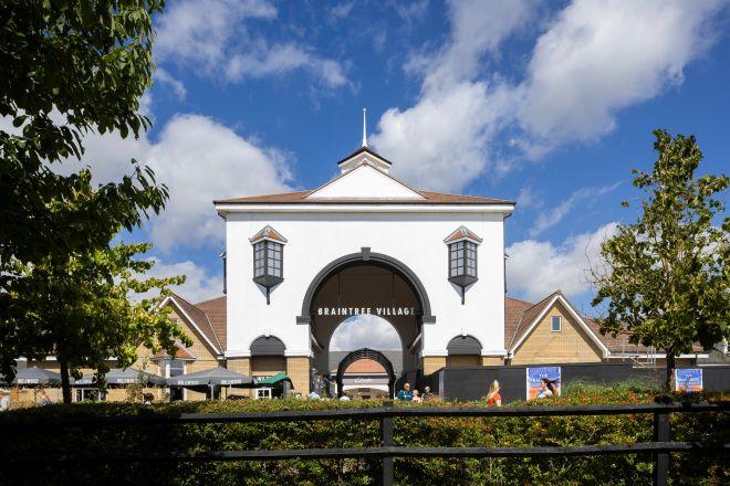 "Main entrance arch at Braintree Village with shoppers walking beneath, framed by trees under a blue sky."