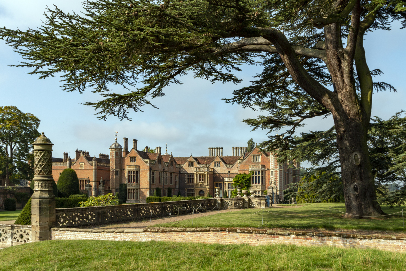 Exteriors of Tudor house at Charlecote Park, Warwickshire. The image features a large building with a spacious lawn in front of it. The outdoor scene includes green grass, a blue sky with clouds, and various trees and plants in the landscaping. The building it built from brick, it has many many windows, chimneys and gables. 