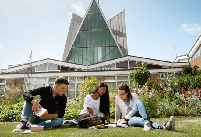 "Image of students sat on the grass outside of an iconic CCCU Building shaped like a pyramid"