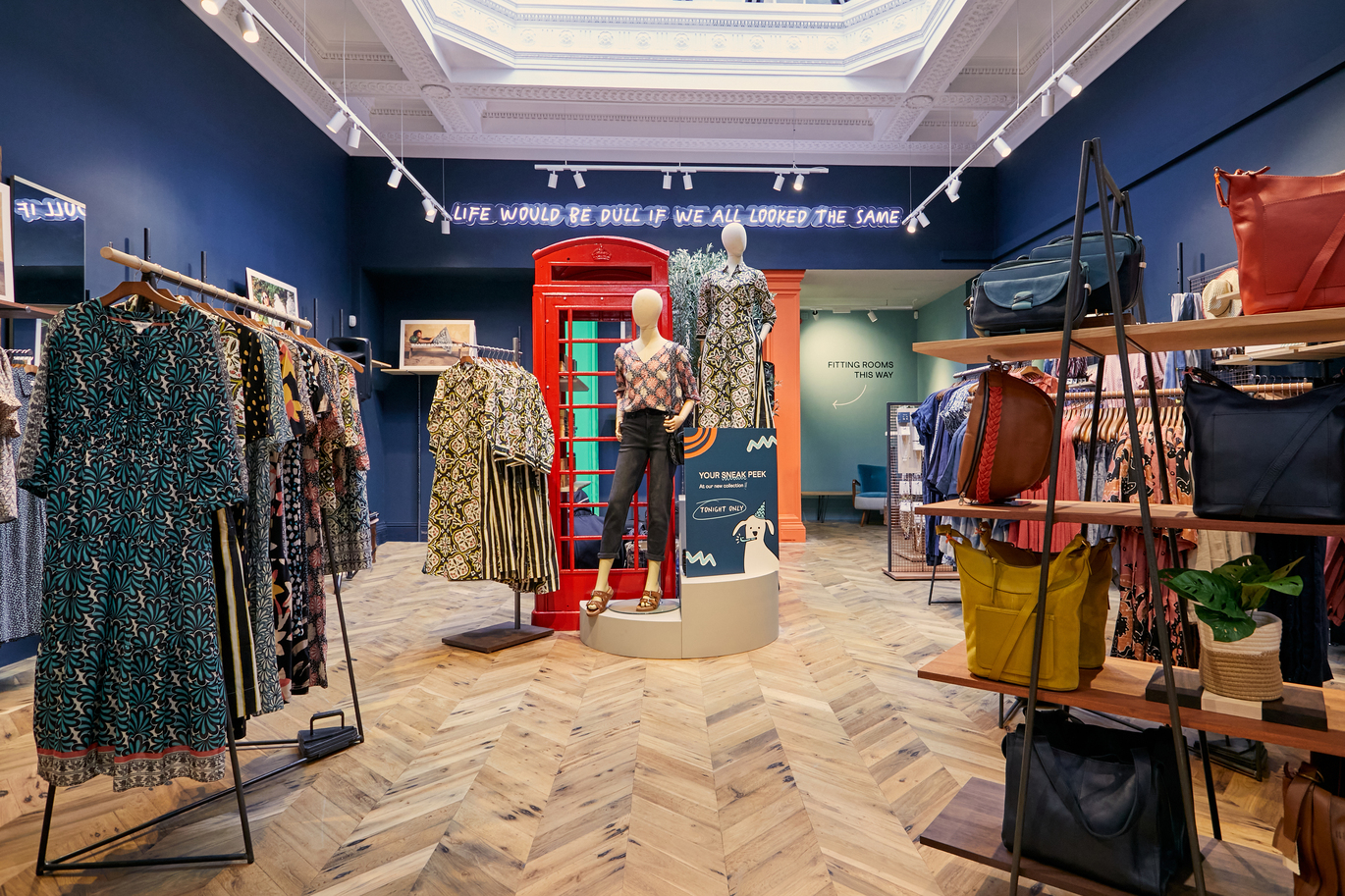 Inside view of a White Stuff store with clothes on rails and bags on shelves, with a couple of mannequins next to a red phone box and wording on the wall up high reading 'Life would be dull if we all looked the same' with fitting rooms right at the back.