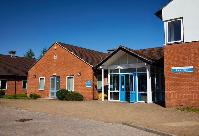 Front view of the brick building with blue front doors and a blue and white sign on the wall that reads 'Reception' with a dropped curb out the front.