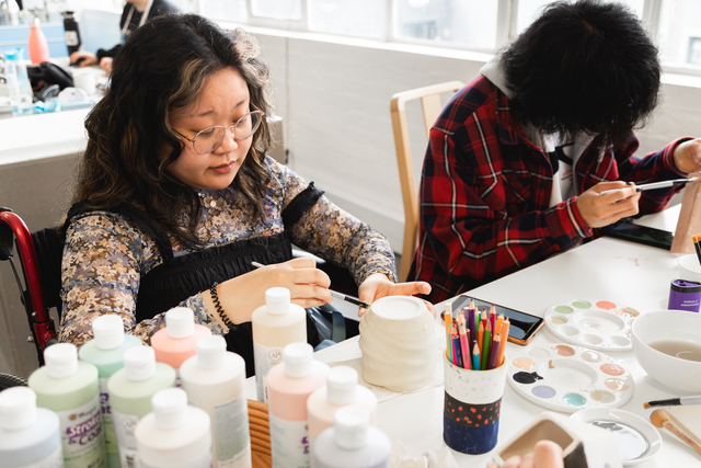 'two people painting pottery, while multiple paint pots in the foreground' 