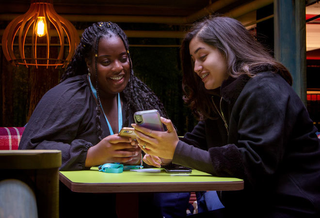two students are sat at a table, laughing at something on one of their phones.