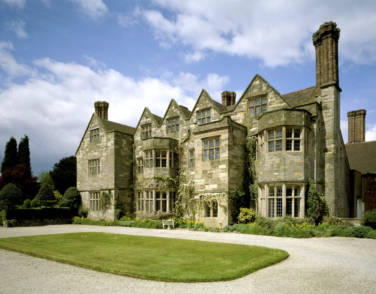 The garden front of Benthall Hall, a 16th century stone house, with a rectangular grassy forecourt and a stone gravel drive around the grass. The building has lots of gables and many different size windows, and a number of chimneys. The building also has climbing plants up the front and lots of greenery around the base. In the background you can see some neatly trimmed hedges of different sizes and shapes. The sky is a lovely blue with white fluffy clouds.