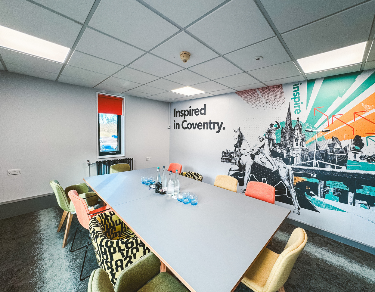 "Small conference room with large table in the middle of the room, chairs surround the table, carpeted floor and mural of Coventry on the wall"