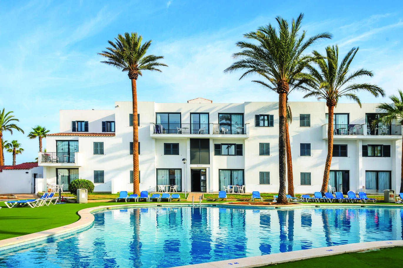 View across the pool of the white hotel building right behind the pool, with blue sun loungers around the pool on the lawns and a few tall palm trees in front of the building, with the blue, slightly cloudy sky above.