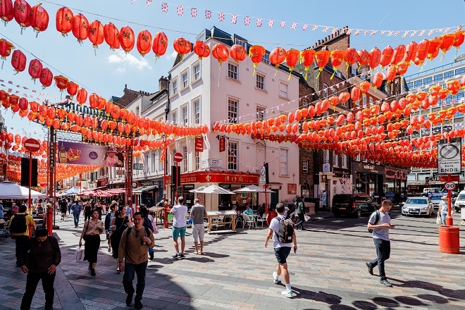"Photo showing view from a corner of Chinatown with shops and restaurants from the left and right. There is a shared road between cars and people, there are lots of red lanterns above going between all buildings."