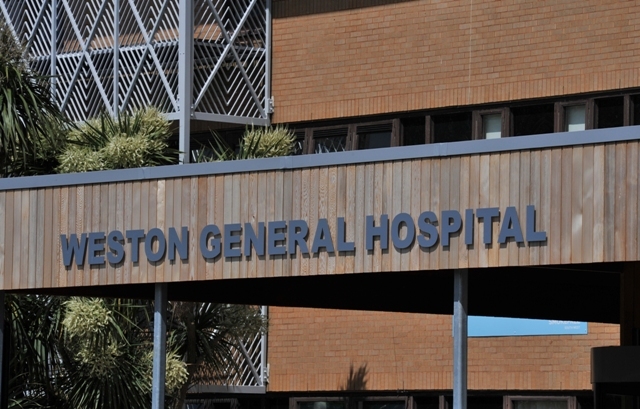 Weston General Hospital front of hospital sign with grey letters on a wooden background with brick and foliage behind.