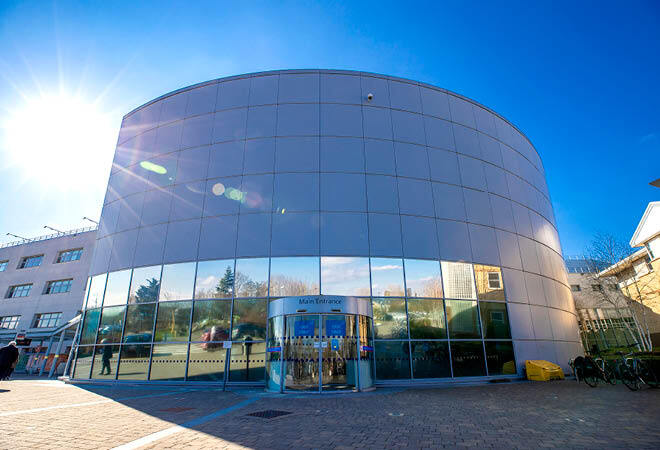 "Main entrance of Broomfield Hospital. Rounded building with mirrored lower section and silver cladding covering the rest of the building. Doorway with sign above saying 'Main Entrance' Blue and sunny skies are visible in the background"