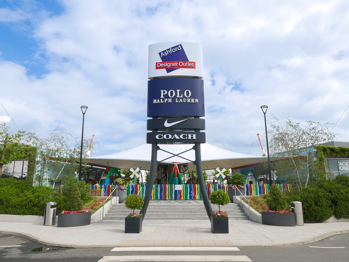 “Entrance of Ashford Designer Outlet featuring a tall sign with brand logos for Polo Ralph Lauren, Nike, and Coach. The sign stands in front of a modern canopy structure with colorful decorations and landscaped planters along the walkway.”