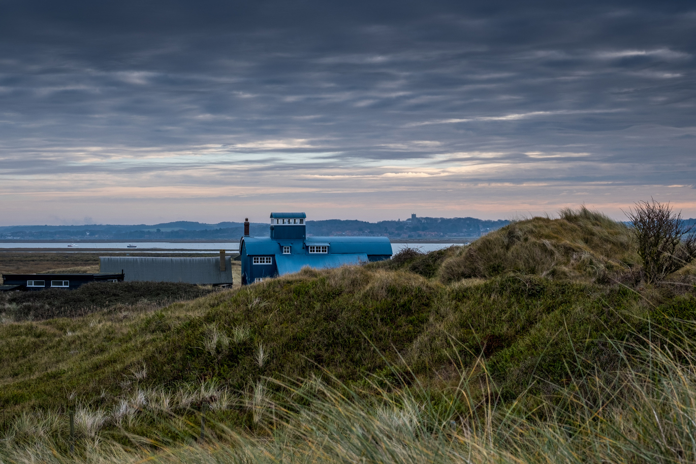 The Old Lifeboat Station at Blakeney National Nature Reserve sitting on top of a grass hill with the sea in the back ground and some boats, and the land beyond on the other side.