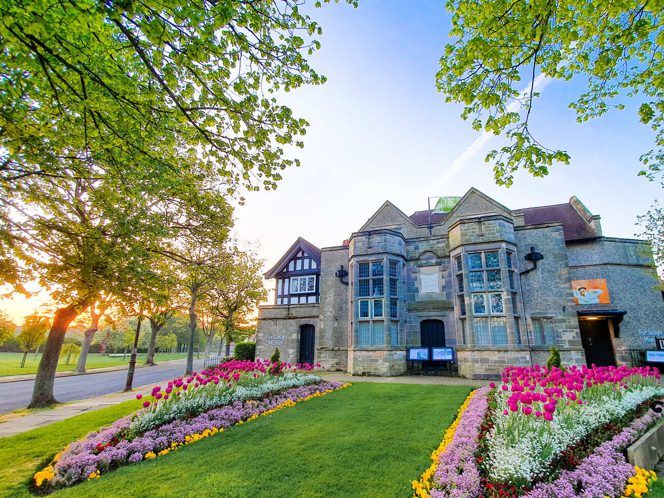 The Port Sunlight Museum, featuring vibrant flower beds and lush greenery, is illuminated by a warm sunset glow.
