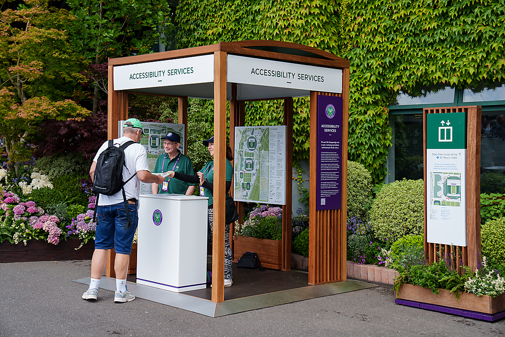 "Image shows the Accessibility Services Booth at Wimbledon with two Wimbledon employees wearing green t-shirts and a member of the public"