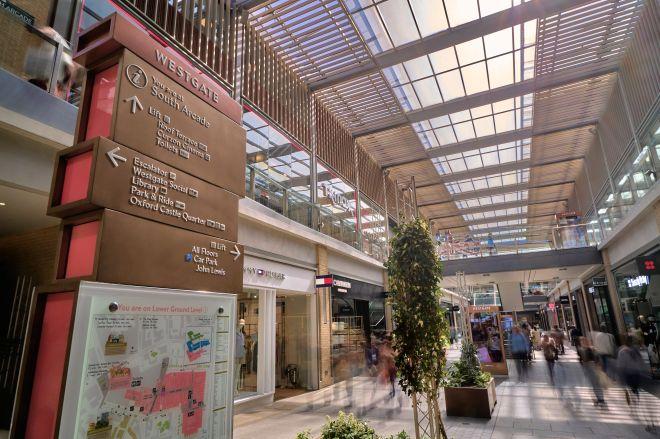 "Indoor shopping centre walkway with glass roof, shops, and a large Westgate directional sign."