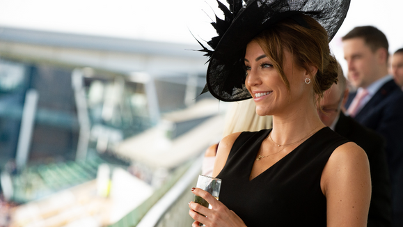 "Lady dressed all in black, with a large black hat drinking champagne in a corporate box"