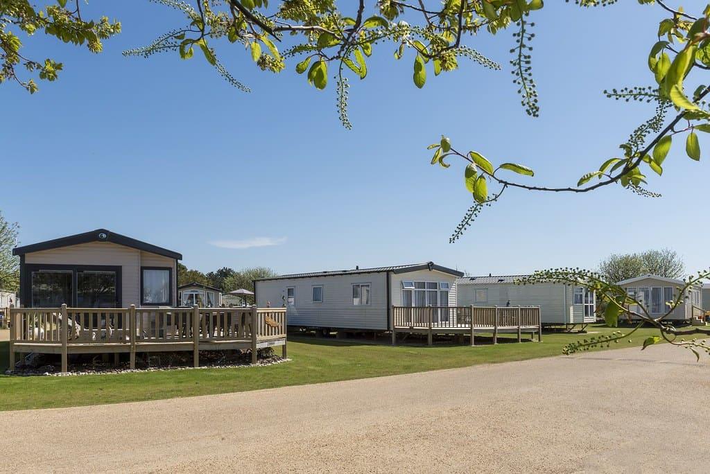A sunny day at a caravan park featuring a row of mobile homes with wooden decks, surrounded by green grass and trees, conveying tranquility and leisure.