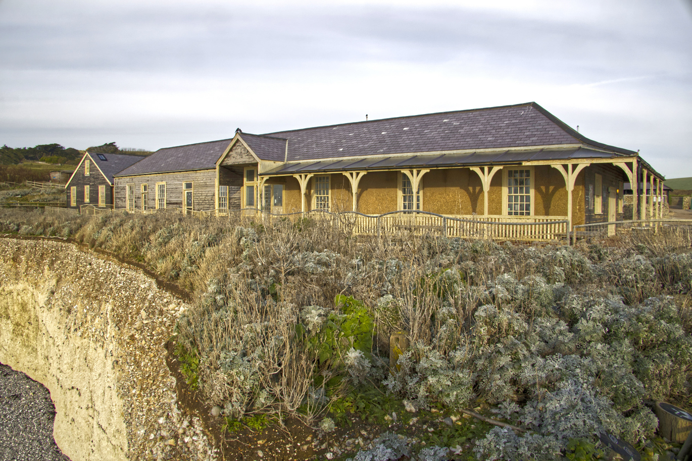 Exterior of the Old Café at the top of a sheer drop to a stony beach surrounded by overgrown greenery on and overcast day. 