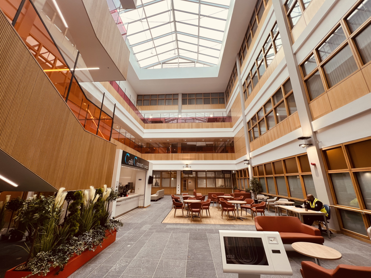 "A café area in an atrium at Ulster Hospital called 'Café Connect'. There are seven circular tables with chairs with armrests around them. A man in a hi vis vest is sat on a desk chair at a desk with a thermal flask in the foreground on the right. He is looking towards the café counter which is on the left of the photo. There is a touch screen pad in the foreground as well as a planter with several plants in. The atrium rises through four floors of the building and there is a large glazed roof where the sky can be seen."