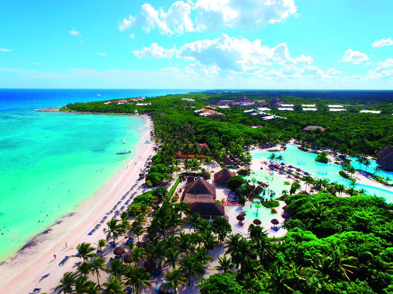 Long aerial shot of the hotel complex with a number of pools around. With many palm trees dotted around the complex and the buildings surrounding. The blue sea is is right next to the complex with beautiful white beaches and the slightly cloudy blue sky above..