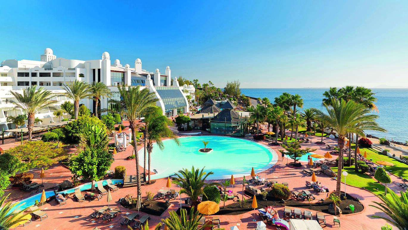 View of the resort with the small round pool in the middle with a small river coming off it with a bridge over it. The large, white hotel building is behind and the sea is just visible over the other side.