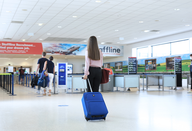 "Photograph shows a person with long brown hair, a pink seater and black trousers, pulling a blue carry on case headed towards security"