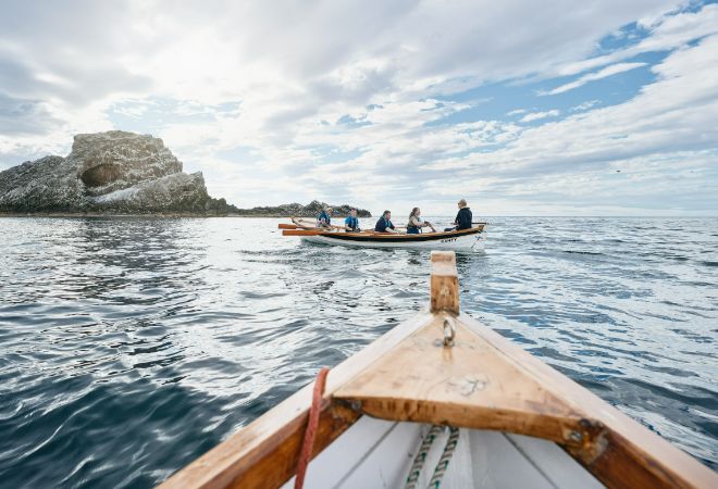 "Cullen Sea School. 5 people in a canoe on the sea in back ground with sunny cloudy skies. Bow of Canoe in foreground. Rock formation in left background."