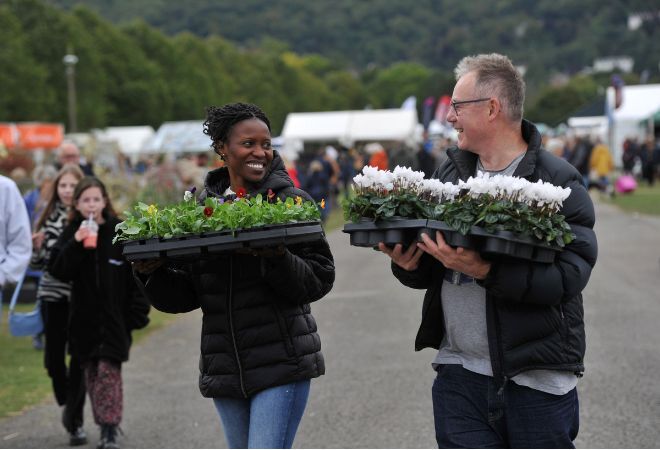 "A femme presenting person and a male presenting person both carrying punnets of flowers at one of the shows" 