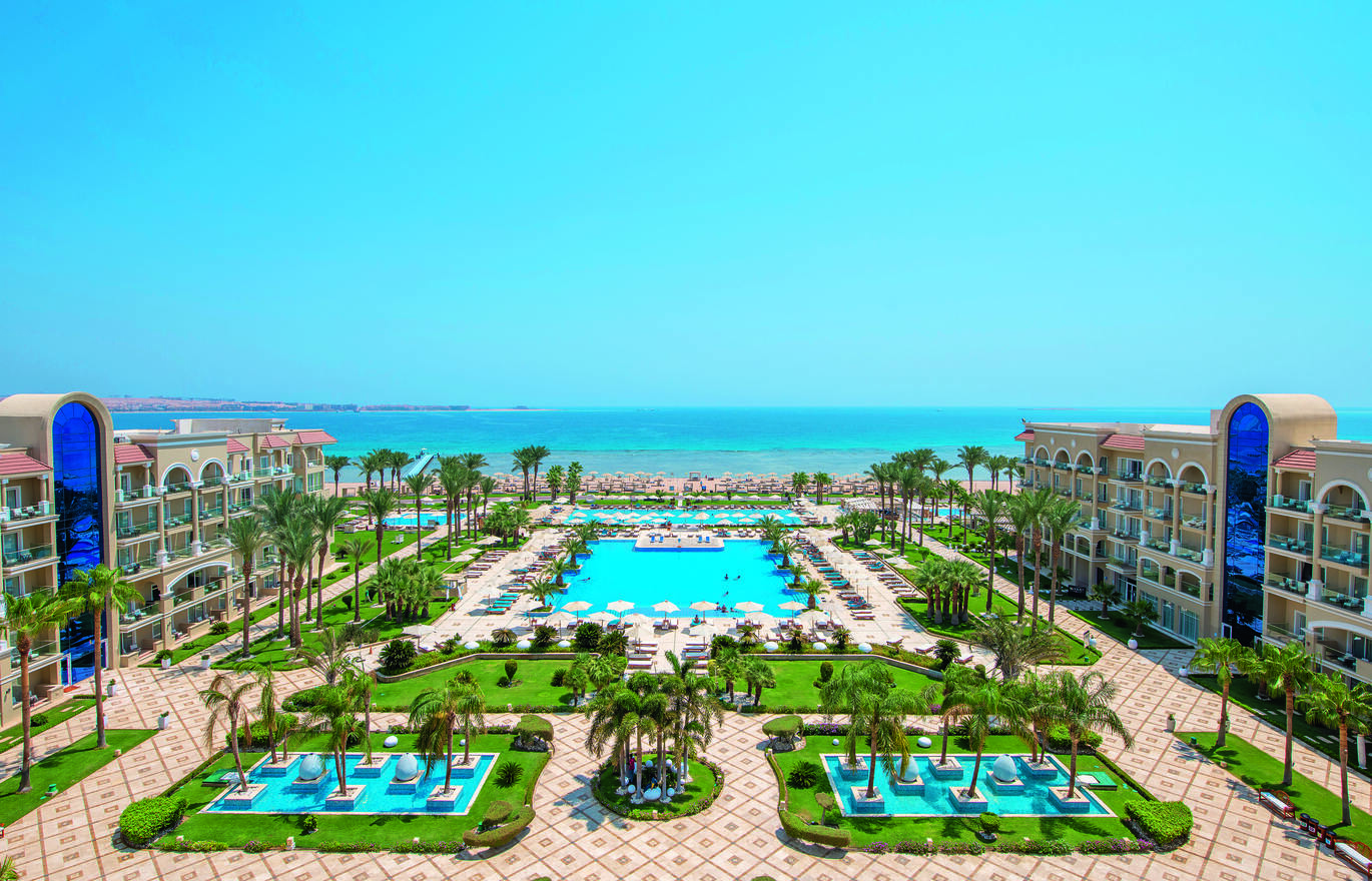 View of the large hotel complex with a number of pools and sun loungers surrounding. With many palm trees around and the hotel buildings either side of the pool. The beach and sea are directly behind.