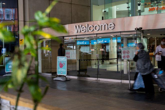 "Shopping centre entrance with glass doors and “Welcome” sign, with people walking past."