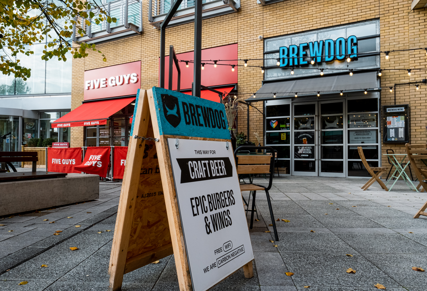"Image shows the entrances to BrewDog and Five Guys from a patio area with seating and tables. 