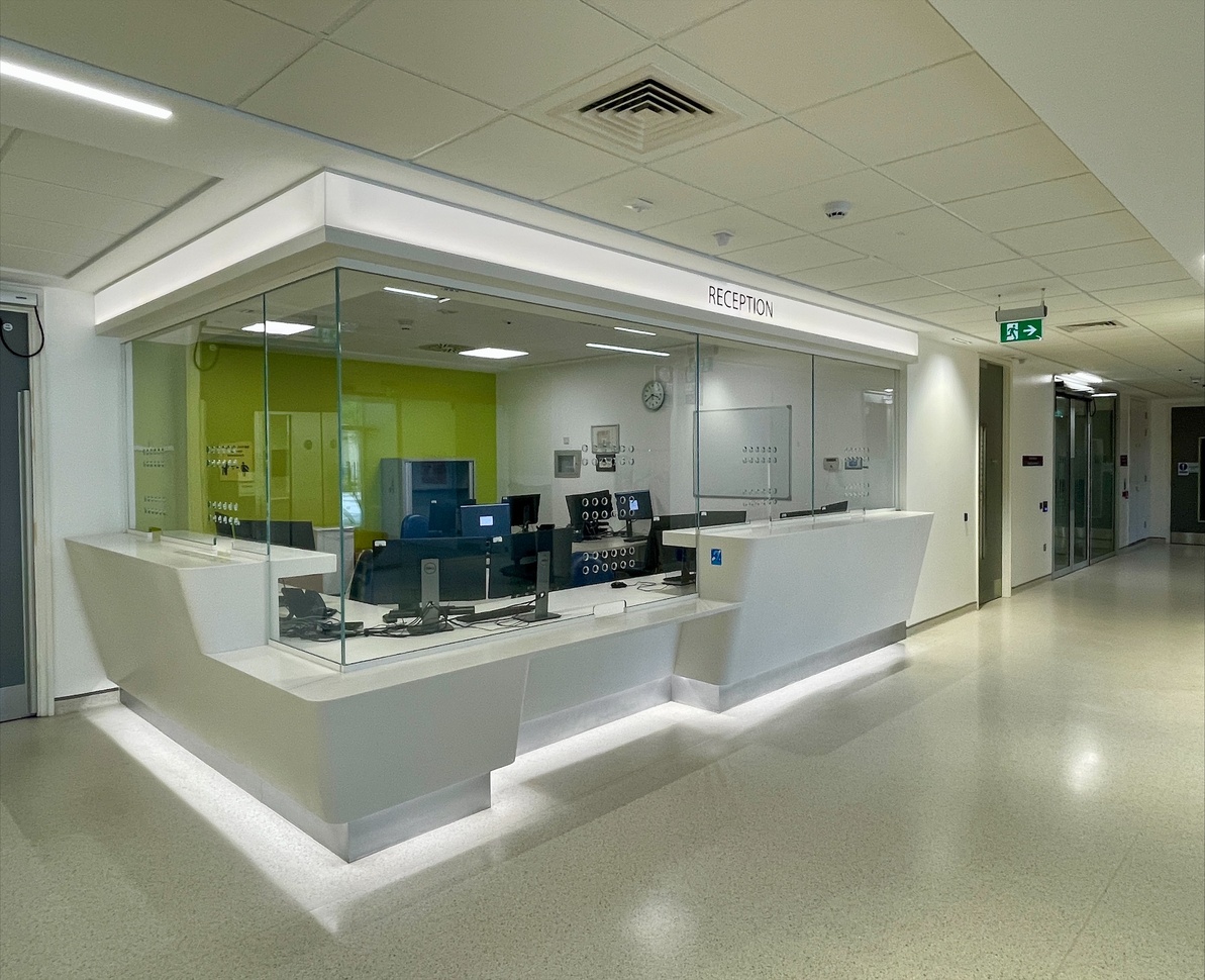 "A hospital reception area enclosed with protective screens. There is a counter with both high and low sections. There are multiple computers at desks in the reception area as well as a whiteboard and clock on the wall."