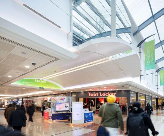 "Indoor shopping centre with high glass roof, Foot Locker store and people walking."