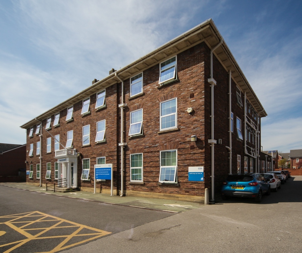 "A close up image of a 3 storey brick built building called Curzon Road Medical Practice. There are NHS signs at the front of the building and a row of cars parked along the side of the building."