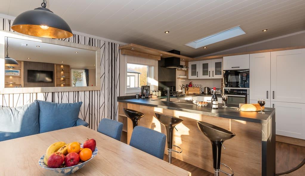 Modern kitchen and dining area with sleek design. Features a wooden island with bar stools, fruit bowl on table, and appliances. Warm, inviting ambiance.