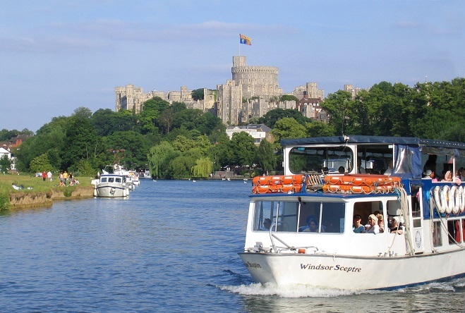 "Photo of river boats on the river, in the background is Windsor Castle with trees in front of it to the side of the river"