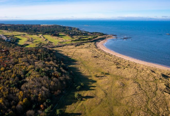 "Aerial view of Yellowcraig Beach. Trees to the left turn into grassland as they approach the beach. The sandy beach curls to the right, and faces the sea."