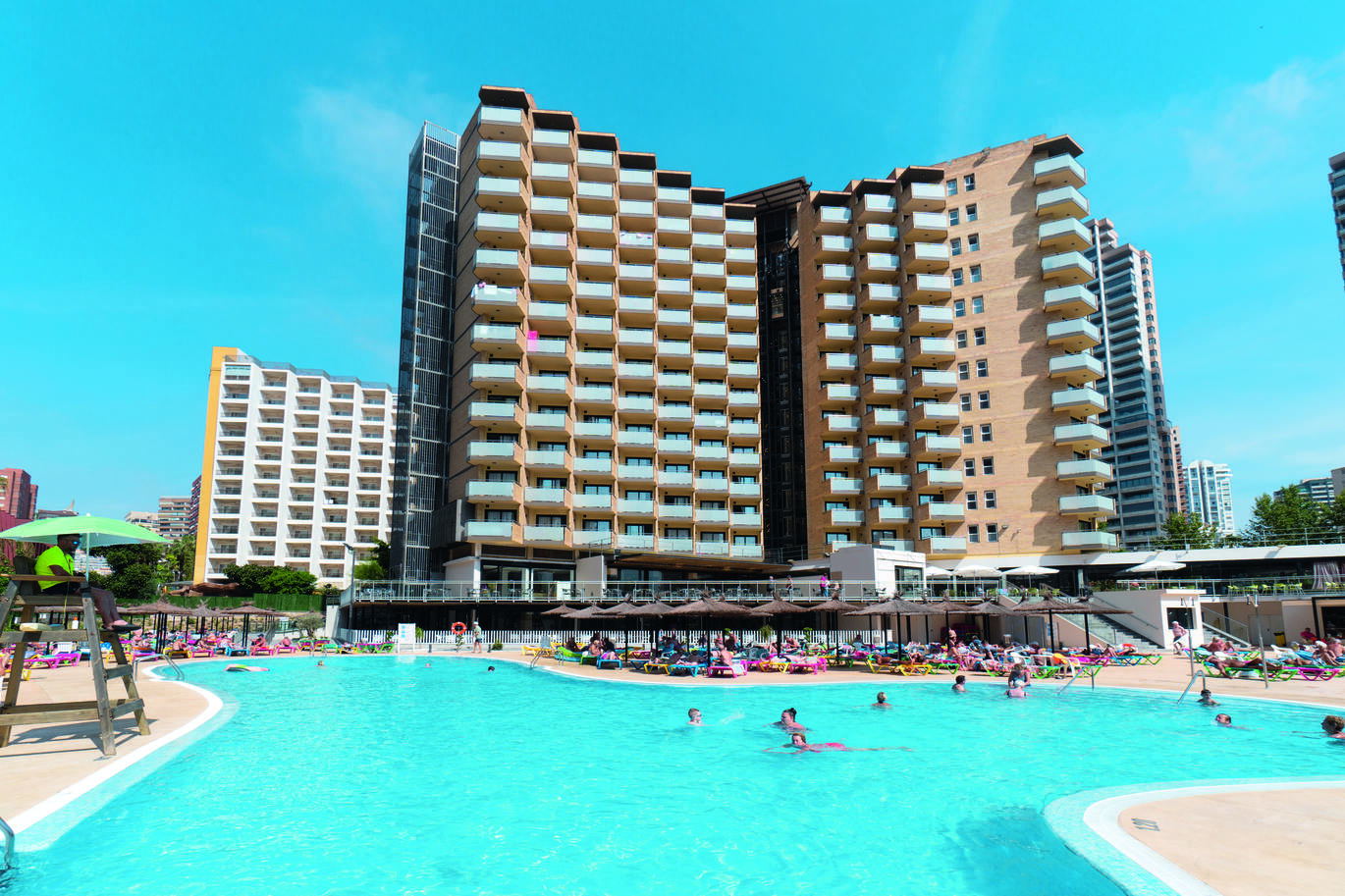 Very tall hotel building in the background with lots of balconies, in front is the blue pool with a number of sun loungers filled with people covered by straw like umbrellas. There is a life guard to the left of the pool and the lovely blue sky above with a few very small clouds.