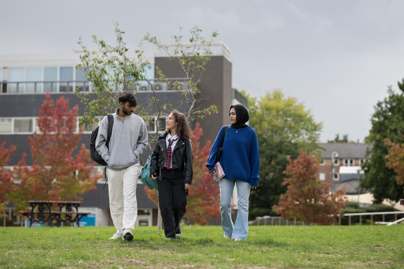 "Three people walk together across a grassy campus area, carrying bags and talking as they move. Modern university buildings and trees with autumn-coloured leaves are visible in the background on a bright day."