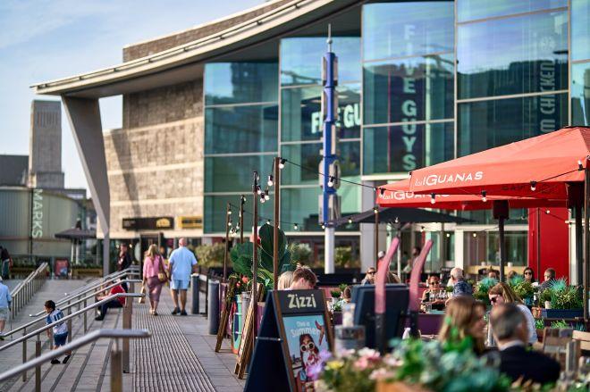 "Waterside dining area at Liverpool ONE with outdoor seating, umbrellas and shoppers walking along the promenade."